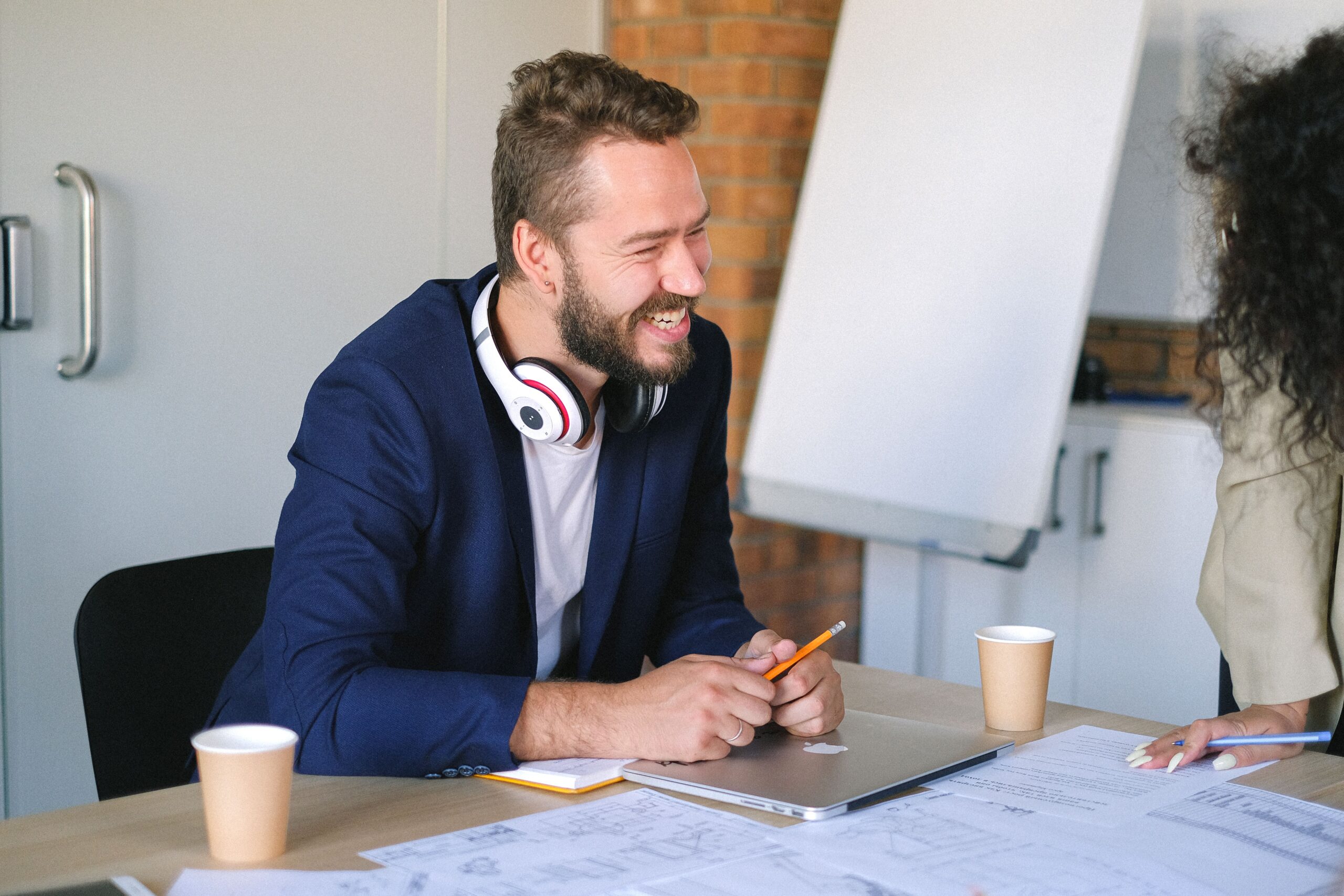Smiling male with headphones sitting at table with documents and laptop while working in office with anonymous colleague